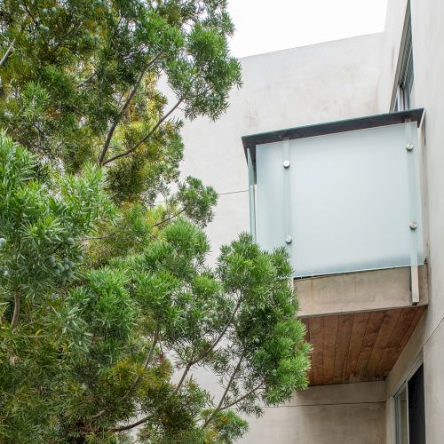 The image shows a small glass-walled balcony on a modern building next to a large green tree.